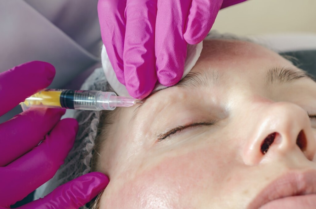 Aesthetic injector administering a facial filler injection near the eye and brow area using a syringe with pink gloves